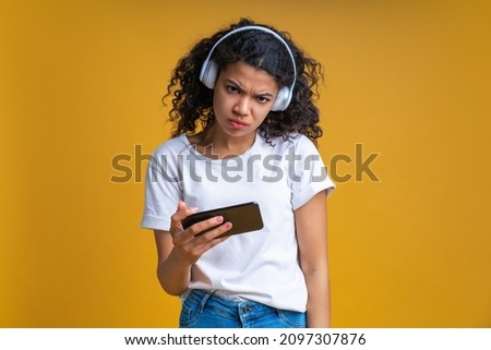 Portrait of frowning girl holding smartphone in hand and looking at the camera with sullen face expression feeling frustrated that she lost online play tournament. Isolated over yellow background.