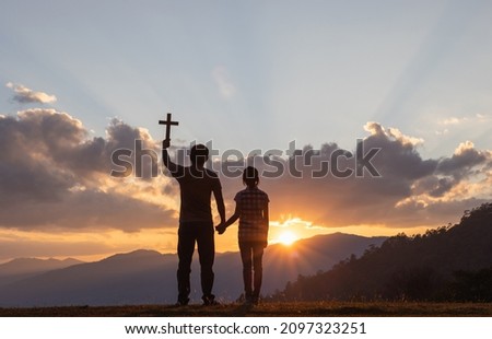 Silhouette Christian father holding Christian cross teaching his daughter to pray outside on mountain sunset background.