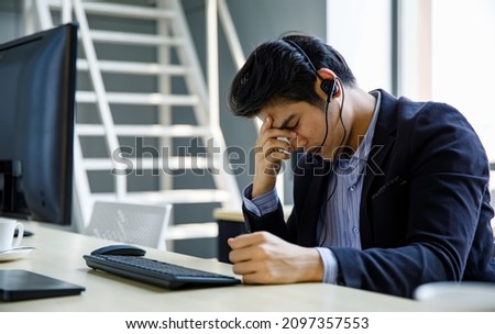 Asian stressed depressed upset telemarketer receptionist operator service support male employee staff in formal suit wears headset holding hand on head having headache when working overload overtime. Royalty-Free Stock Photo #2097357553