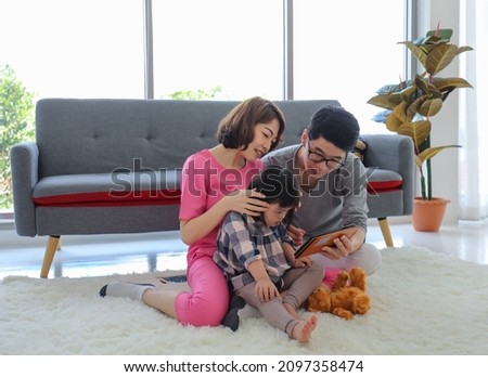 Overjoyed happy family Asian father and mother sitting on carpet floor holding touchscreen tablet computer showing to little cute daughter watching cartoon together in living room at home on weekend.