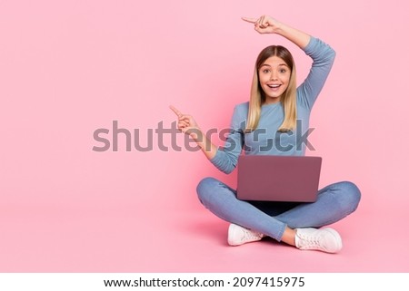 Full size photo of young girl point fingers empty space promo recommend direct sit floor laptop isolated over pink color background