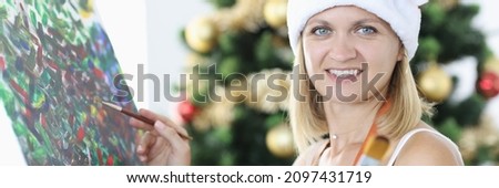 Smiling woman artist in santa claus hat draws picture against background of Christmas tree. Profession artist and Christmas concept