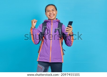 Portrait of excited young Asian man celebrating with mobile phone isolated on blue background