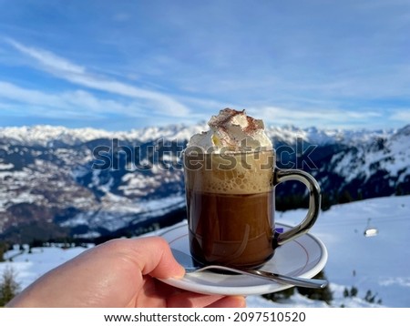 Apres ski, after ski in the Austrian Alps. Hand holding cup of hot chocolate with whipped cream, snow covered mountains in the background.