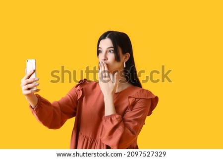 Surprised young woman with mobile phone taking selfie on yellow background