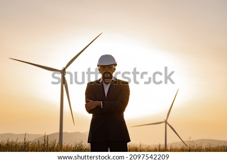 Indian businessman in black suit, white helmet and safety glasses posing with crossed arms among field with wind turbines. Amazing summer sunset on background.