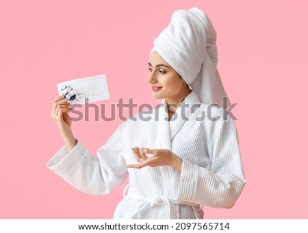 Pretty young woman in bathrobe showing gift voucher on pink background