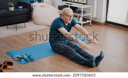 Retired man doing physical exercise to practice gymnastics and fitness activity. Senior person stretching arms and legs for healthy workout, sitting on yoga mat. Active pensioner