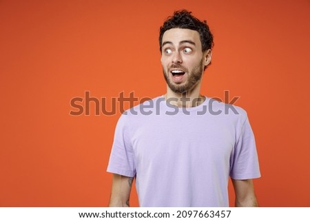 Shocked amazed surprised excited young bearded man 20s wearing basic casual violet t-shirt standing keeping mouth open looking aside isolated on bright orange color wall background studio portrait