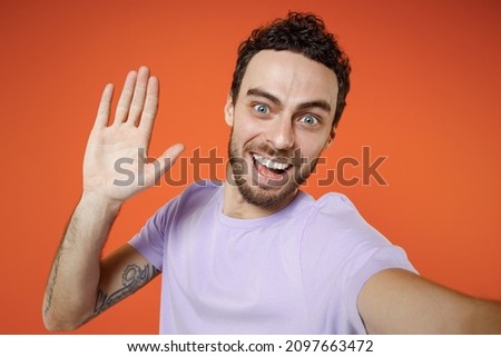 Close up of excited young bearded man 20s wearing casual violet t-shirt standing doing selfie shot on mobile phone greeting with hand isolated on orange background studio portrait Tattoo translate fun