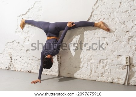 A woman trainer practicing yoga performs Samakonasana exercise in adho mukha vrikshasana, stands on one hand with a transverse twine leaning against the wall, an inverted asana