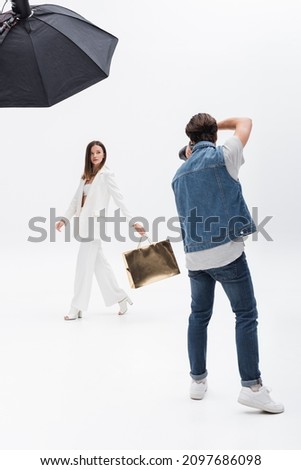 back view of photographer taking photo of model in white suit walking with golden shopping bag on white