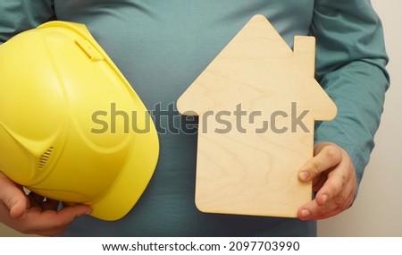 builder man holding in hands yellow helmet and wooden sign of house. Isolated on white background. 