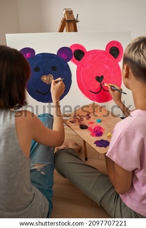 Back view of young lesbian girls painting picture of mouse faces with paints on easel at home. Domestic entertainment, hobby and leisure. Homosexual relationship and spending time together. Daytime