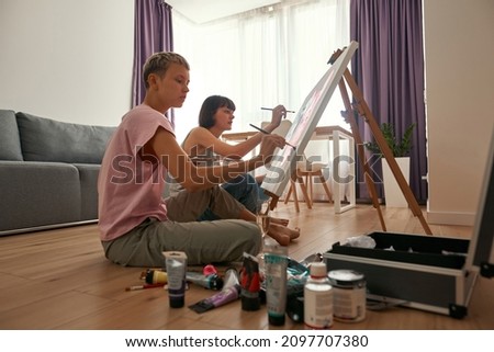 Young concentrated lesbian caucasian girls painting picture of mouse faces with paints on easel at home. Domestic entertainment, hobby and leisure. Homosexual relationship and spending time together