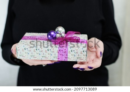 Female hands with long nails and bright purple and blue manicure hold a gift box and Christmas balls. Congratulations to the New Year and Christmas.