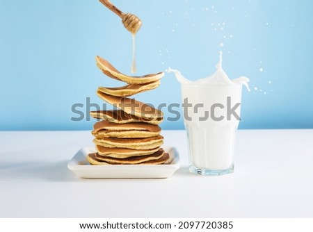 Shrove Tuesday. Levitating stack of pancakes with honey and glass of milk. Splash of milk from the glass on a blue background.  Levitation, gravity. Homemade breakfast. Royalty-Free Stock Photo #2097720385