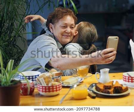 Grandma and granddaughter are sitting in the kitchen, looking at photos on their phones and taking selfies. Warm family relationships. Joy of communication.
