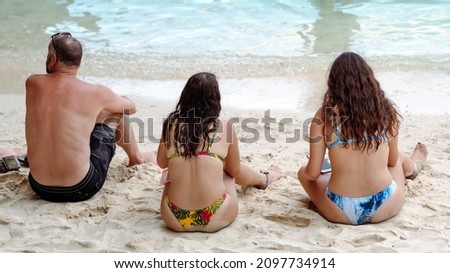 Female caucasian tourist in bikini using her smartphone and sunbath while relaxing with friends on tropical beach.Happy summer holidays and travel vacation.Cheerful woman enjoying summer day at beach.