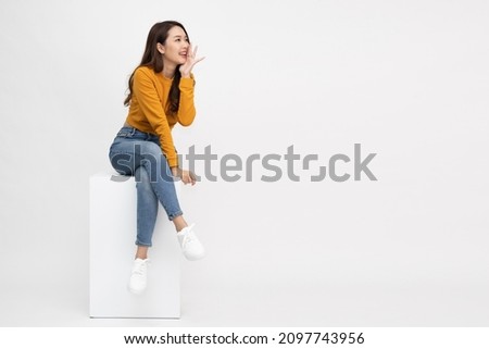 Portrait of young Asian woman sitting on white box and keeps hand near mouth and whispers secret isolated over white background, Spreads rumors concept Royalty-Free Stock Photo #2097743956