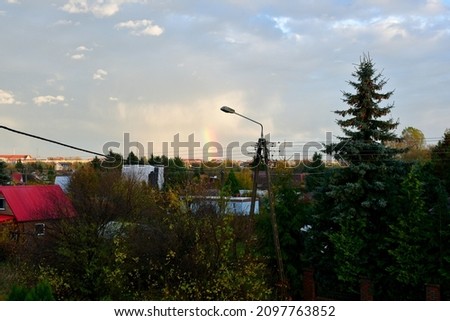 A view of a small rainbow in the clouds seen from the roof of a tall building, also showing a vast village, some trees and a big concrete lamp spotted on a Polish countryside in summer