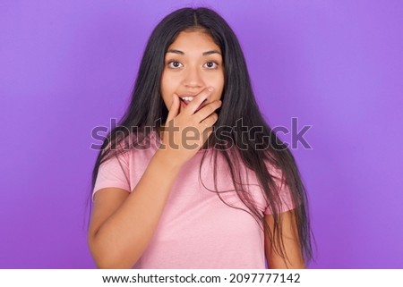 Emotional Hispanic brunette girl wearing pink t-shirt over purple background gasps from astonishment, covers opened mouth with palm, looks shocked at camera. Reaction concept