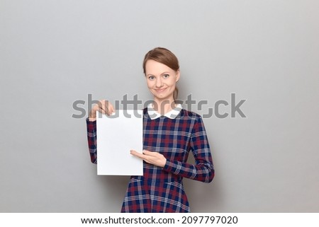 Studio portrait of happy young woman holding white blank paper sheet, pointing with palm at empty place for your text and design, smiling, wearing checkered dress, standing over gray background