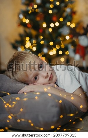 Portrait of a smiling cute boy 5-7 years old lying on the floor against the background of a Christmas tree. Selective soft focus. blond boy near the Christmas tree lying on the floor. New Year