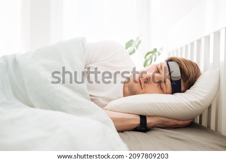 Man sleeping on bed with smart sleep headband. Smart sleep tracker. Heartbeat monitor on head. Modern male health monitor. Cozy sleeping bed room. Wearable technology in a daily life to develop habit Royalty-Free Stock Photo #2097809203