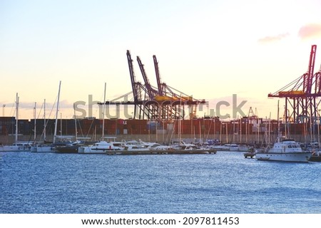 Container crane in seaport on sunset. Yachts and motor boats in La Marina de Valencia. Unloading sea martime containers. Container ship and  shipping containers box. Luxury yacht and Sailboat in port.
