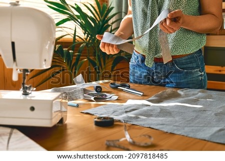 Woman cutting out a pattern paper in linen fabric. Seamstress sewing on the sewing machine in small studio. Fashion atelier, tailoring, handmade clothes concept. Slow Fashion Conscious consumption