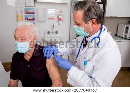 A White Male Medical Doctor Administering a Covid-19, SARS-CoV-2 Vaccine Injection with a Syringe Needle to an Elderly Senior Male Patient Wearing Gloves and Mask in Hospital or Health Clinic.