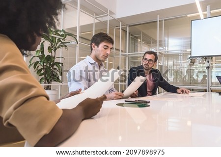3 mixed hispanic latin business people, men and woman, sitting at a table meeting inside an insurance office reading a contract that they must sign with a lawyer watching them.