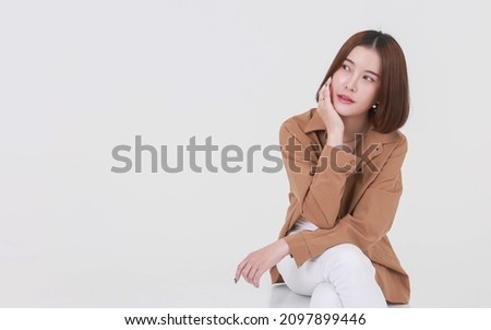 Portrait studio cutout shot of Asian young pretty short hair female model in long brown coat jacket casual shoes sitting crossed legs smiling look at camera on box chair posing on white background.