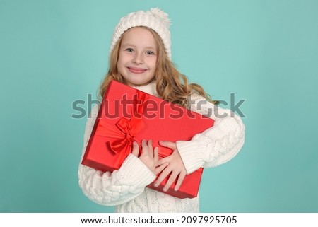 a beautiful blonde girl in a knitted white sweater and a winter hat holds a red gift box in her hands