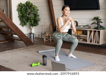 Young asian fitness girl doing squats workout at home, follow online sport gym instructor, standing on floor mat and smiling, exercising in living room Royalty-Free Stock Photo #2097956893