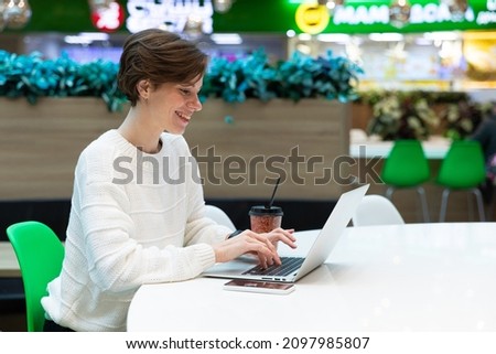 Photo of a young positive woman who sits at the food court of a shopping center at a table and works at a computer laptop. Freelance concept