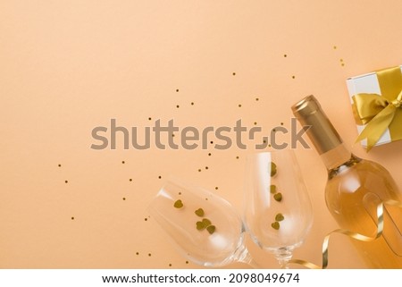 Top view photo of valentine's day decorations white giftbox with golden bow wine bottle two wineglasses with heart shaped confetti and sequins on isolated pastel beige background with empty space