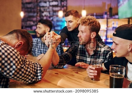 Friends plays arm wrestling. Group of people together indoors in the pub have fun at weekend time.
