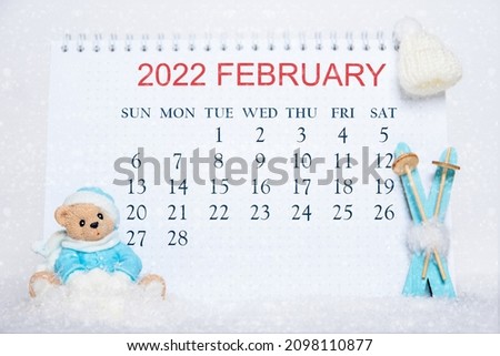 Calendar for the month of February 2022. Notebook with calendar dates and a toy Teddy bear, blue skis, white hat on snow