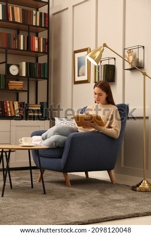 Young woman reading book in armchair indoors. Home library Royalty-Free Stock Photo #2098120048