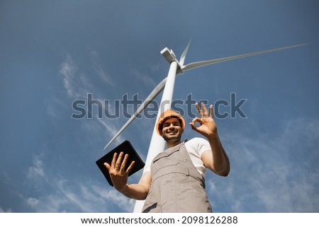Competent indian technician smiling and showing sign OK wind turbines on background. Man in helmet and overalls standing at rural area and holding modern tablet.