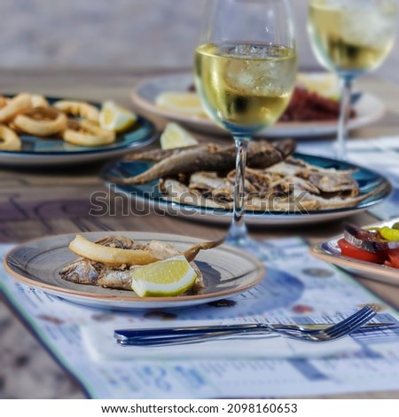 A table with seafood and two glasses of white wine with ice. Squid, fried fish, salad with salted dried tuna, octopus on the background of a sandy beach.
