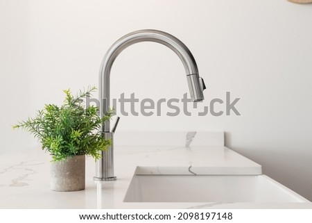 A kitchen sink detail shot with a farmhouse sink, stainless steel faucet, marble countertops, and a plant.