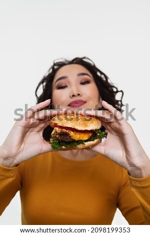 A curly smiling Asian girl in a mustard long sleeve looking at the burger she’s holding with two hands in front of her