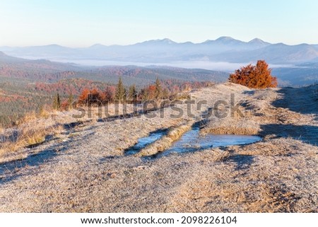 Autumn mountain landscape, frozen road with a frozen puddle and hoarfrost, trees with red foliage, foggy mountains on the horizon.