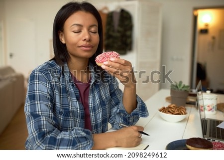 Indoor picture of dark-skinned woman holding pink glazed doughnut with eyes closed enjoying its smell, while having coffee break during work from home, sitting at kitchen table in front of laptop