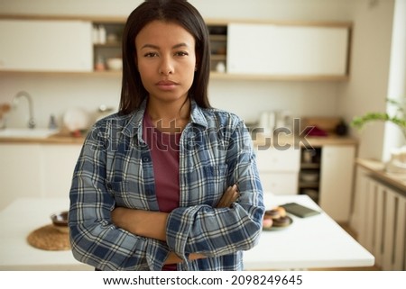 Suspicious mind. Beautiful middle-aged dark-skinned woman in blue plaid shirt standing with arms folded at kitchen looking at camera with mistrust, anger and grudge. Negative human emotions Royalty-Free Stock Photo #2098249645