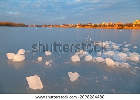 Winter river with ice-hole at sunset, view of the village houses. Soft pink light.