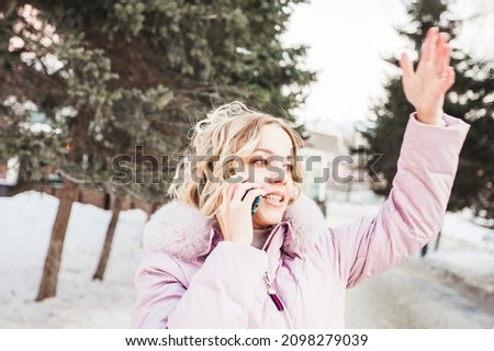 on a winter city street, a young woman waves to someone and talks on a mobile phone